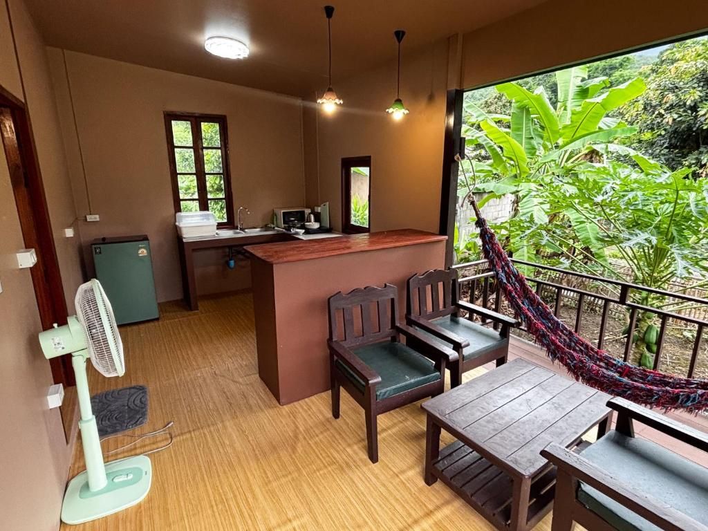 A room with a wooden table and chairs, a small kitchenette, and a view of lush greenery through a large window.