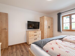 Une chambre à coucher avec une armoire en bois, une télévision, un lit recouvert d’une couverture à carreaux et une fenêtre avec des rideaux.