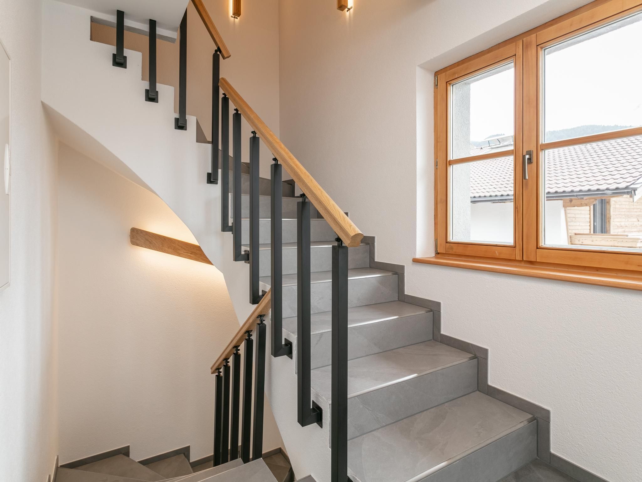 Un escalier moderne avec une main courante en bois et des balustres noires, menant à un palier doté d’une grande fenêtre.