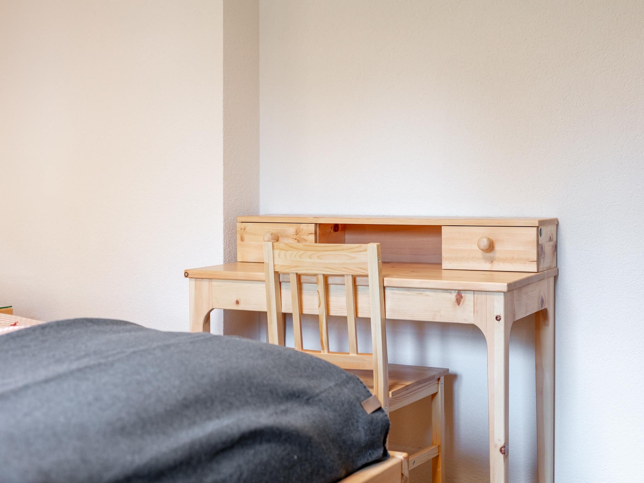 Ein Holztisch mit einem Stuhl, ein Bett mit grauer Decke und eine weiße Wand.
