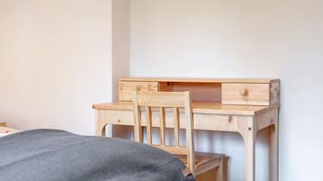Ein Holztisch mit einem Stuhl, ein Bett mit grauer Decke und eine weiße Wand.