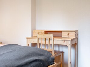 Ein Holztisch mit einem Stuhl, ein Bett mit grauer Decke und eine weiße Wand.