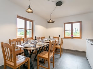 Ein Essbereich mit einem runden Tisch, gedeckt für vier Personen, umgeben von Holzstühlen. Zwei Pendelleuchten befinden sich über dem Tisch und ein Fenster bietet Blick auf Grünflächen.