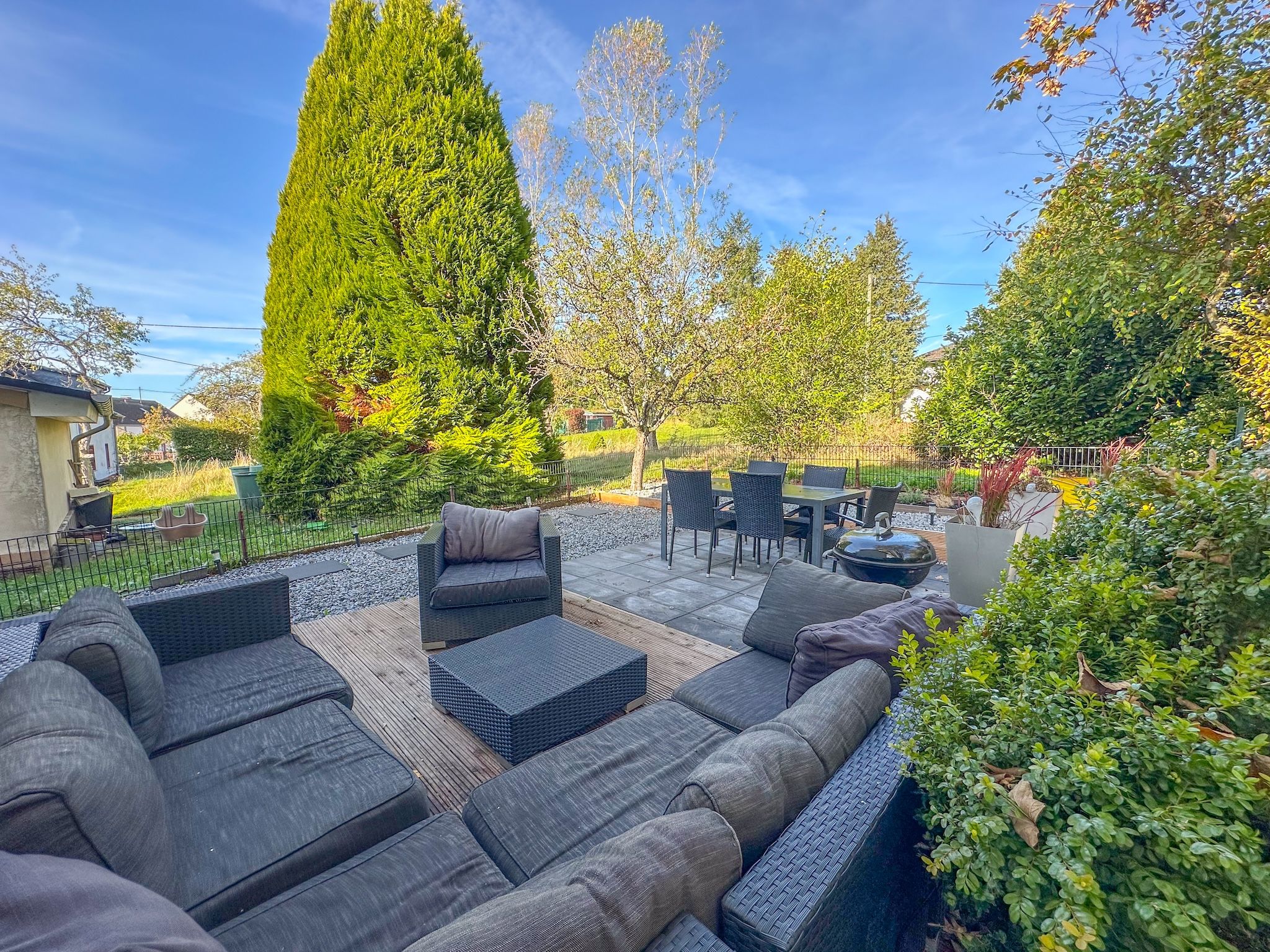 Une terrasse avec des meubles d’extérieur, un grand arbre et une clôture.