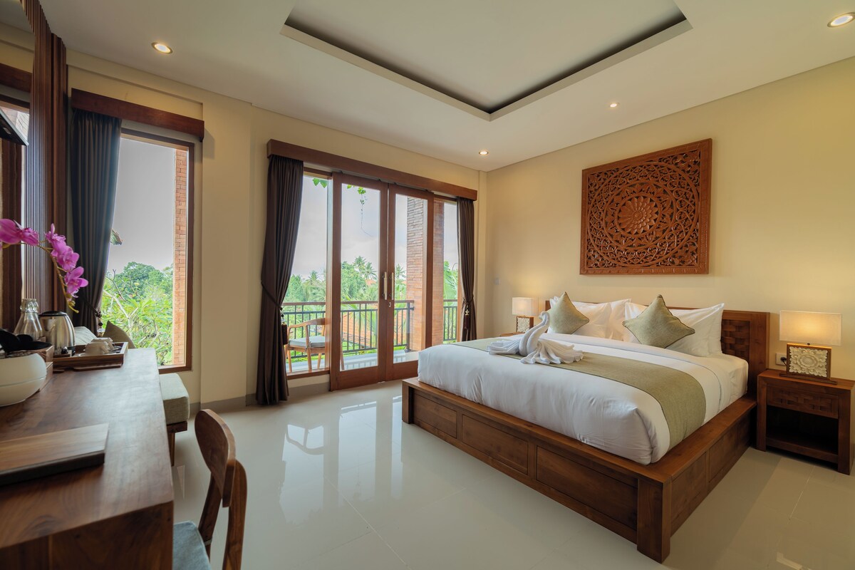 A hotel room with a large bed, a dining table, and a balcony with a view of greenery.