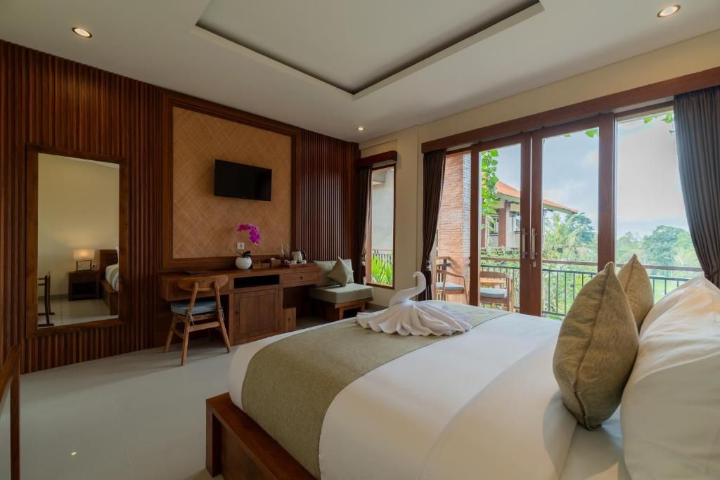 A hotel room with a large bed, a desk, a chair, a TV mounted on the wall, and a balcony with a view of greenery.