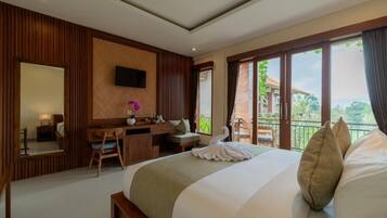 A hotel room with a large bed, a desk, a chair, a TV mounted on the wall, and a balcony with a view of greenery.