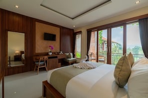A hotel room with a large bed, a desk, a chair, a TV mounted on the wall, and a balcony with a view of greenery. - Donald Ubud Cottages (Ubud)