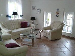 A living room with a sofa, armchair, and coffee table. There are two lamps, a window with curtains, and a door. - Holiday Apartment With 1 Bedroom (Krummhoern)