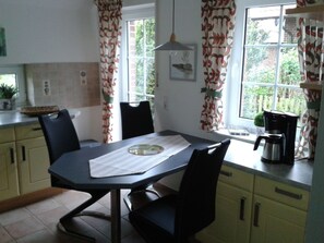 A kitchen with a dining table, chairs, a coffee maker, and a window with curtains. - Holiday Apartment With 1 Bedroom (Krummhoern)