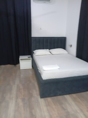 Un llit individual amb matalàs i almoines blanques, una taula de nit i aire condicionat fixat a la paret.