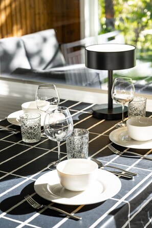 Une table à manger dressée avec des assiettes, des bols, des verres et des couverts, recouverte d’une nappe à motifs et agrémentée d’une lampe en arrière-plan.