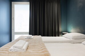 Ein Hotelzimmer mit zwei Betten, einem Fenster mit Jalousien, einem dunklen Vorhang und einem Nachttisch mit Lampe.