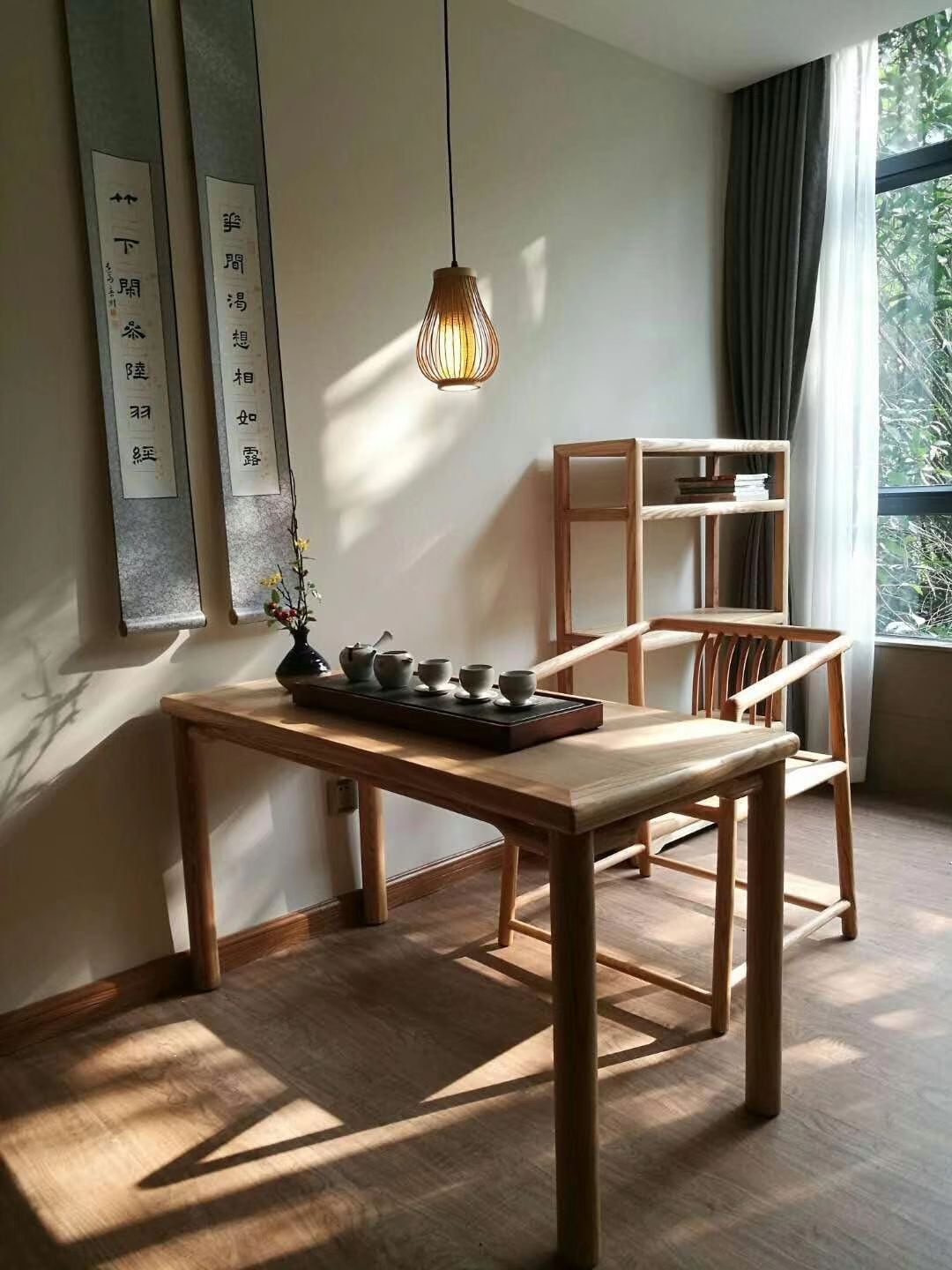 Una habitació amb taula de fusta, llum penjant, prestatges i finestra amb cortines.
