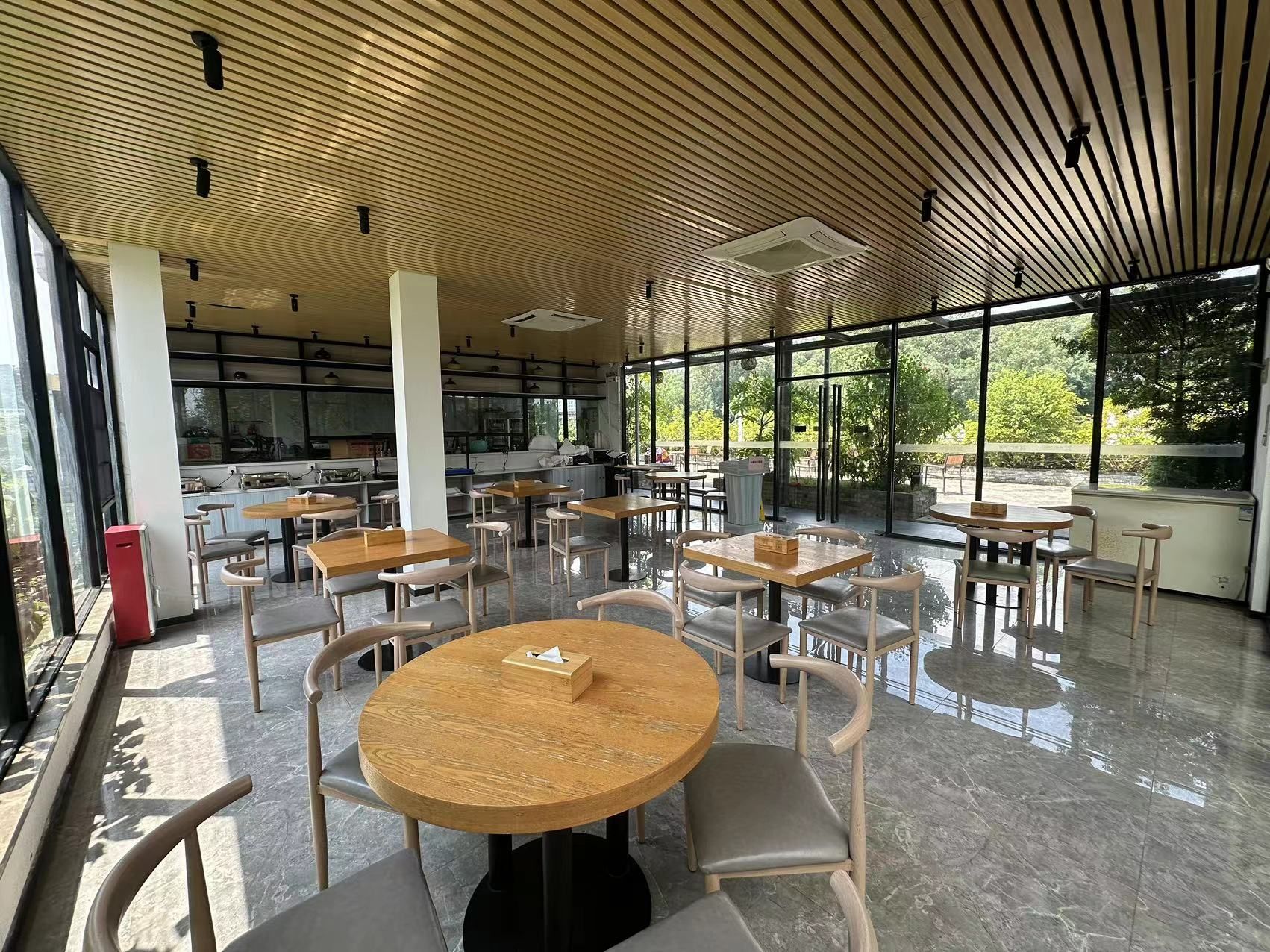 A spacious indoor area with multiple round tables and chairs, large windows offering views of greenery, and a kitchen area in the background.