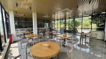 Un espacio interior amplio con varias mesas y sillas redondas, grandes ventanales que ofrecen vistas a zonas verdes, y una cocina al fondo.