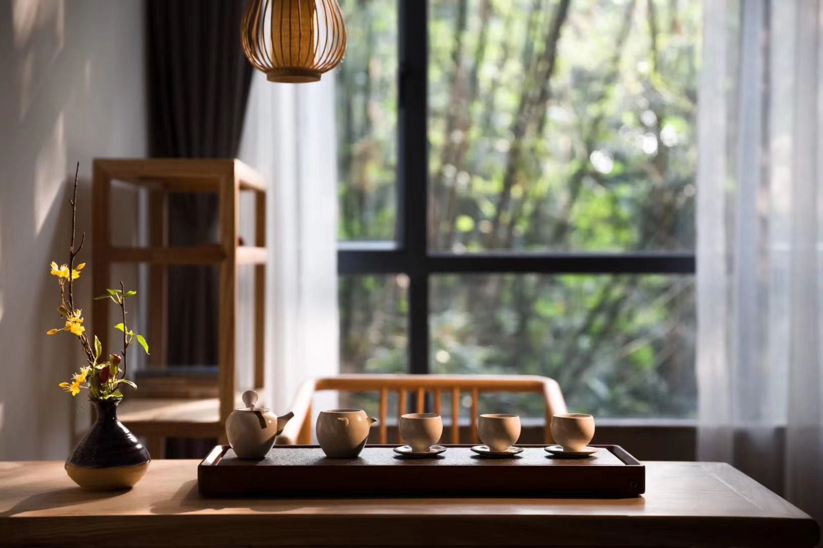 A wooden table with a tray holding tea cups and a vase with flowers, positioned near a window with a view of trees.