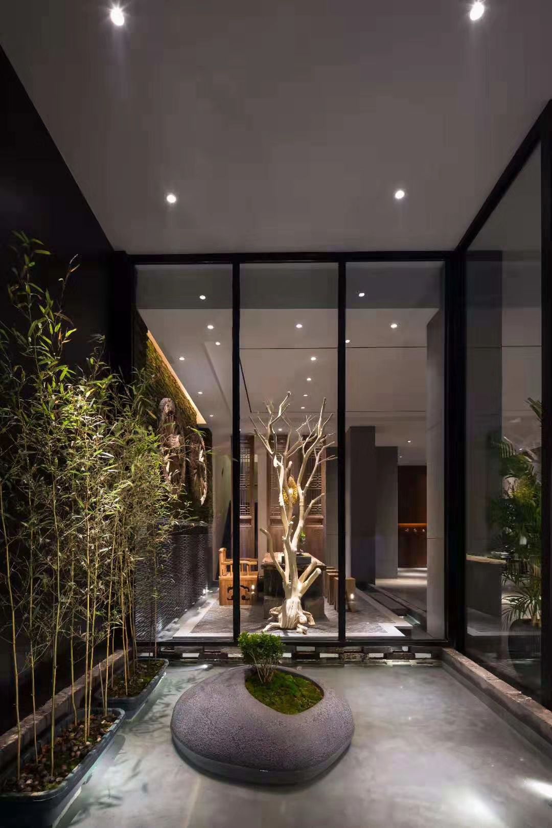 A modern indoor space with a central tree sculpture, glass walls, and a seating area with tables and chairs.