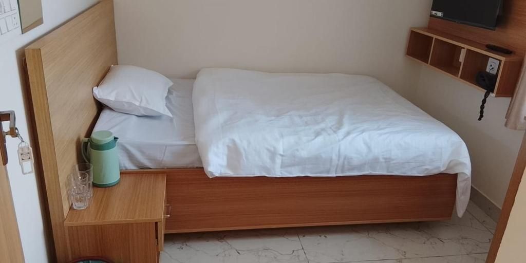 Ein Einzelbett mit Holz Kopfteil, weißer Bettwäsche, einem Nachttisch mit einer grünen Wasserflasche und einem an der Wand befestigten Fernseher.