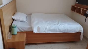Ein Einzelbett mit Holz Kopfteil, weißer Bettwäsche, einem Nachttisch mit einer grünen Wasserflasche und einem an der Wand befestigten Fernseher.