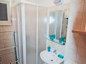A bathroom with a glass shower enclosure, a sink with a mirror above it, and a shelf with toiletries. - Beachfront Apartment With 1 Bedroom (Posedarje)