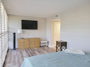 Ein Schlafzimmer mit einem Bett, einem an der Wand befestigten Fernseher, einer Kommode, einem Nachttisch und einer Tür zu einem anderen Raum.