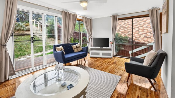 Amplia sala de estar con un gran ventanal que da a un jardín, un sofá azul, un sillón negro, una mesa de centro de vidrio y un televisor de pantalla plana.