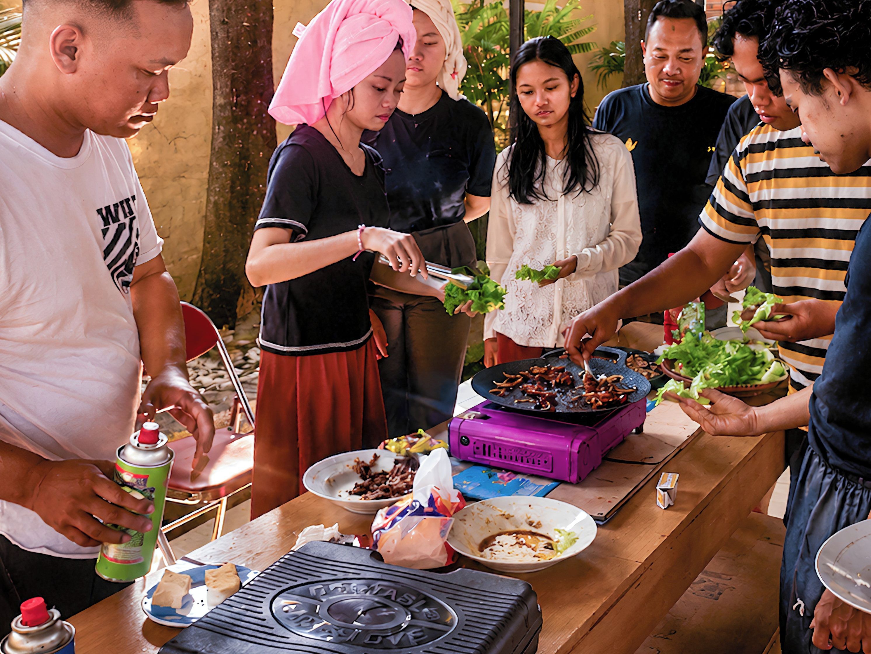 A group of people around a table with food, a portable stove, and a grill.