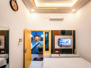 A hotel room with a bed, a TV mounted on the wall, a clock, and an air conditioning unit.