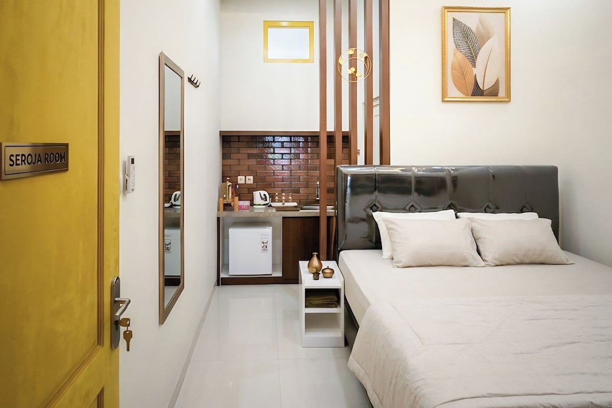 A hotel room with a bed, a small bedside table, a mirror, a wall-mounted picture, a door with a sign, and a kitchenette area.