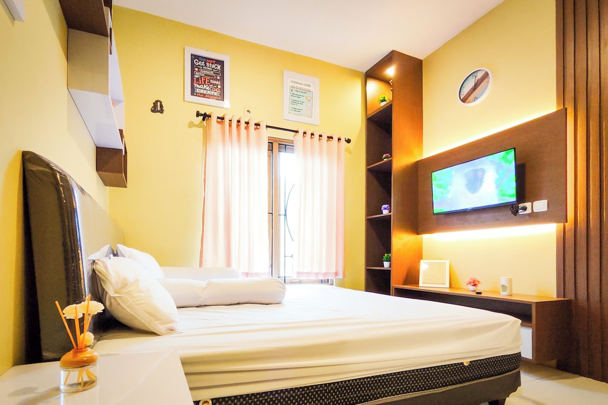 A hotel room with a bed, a television, a shelf with decorative items, and a window with curtains.