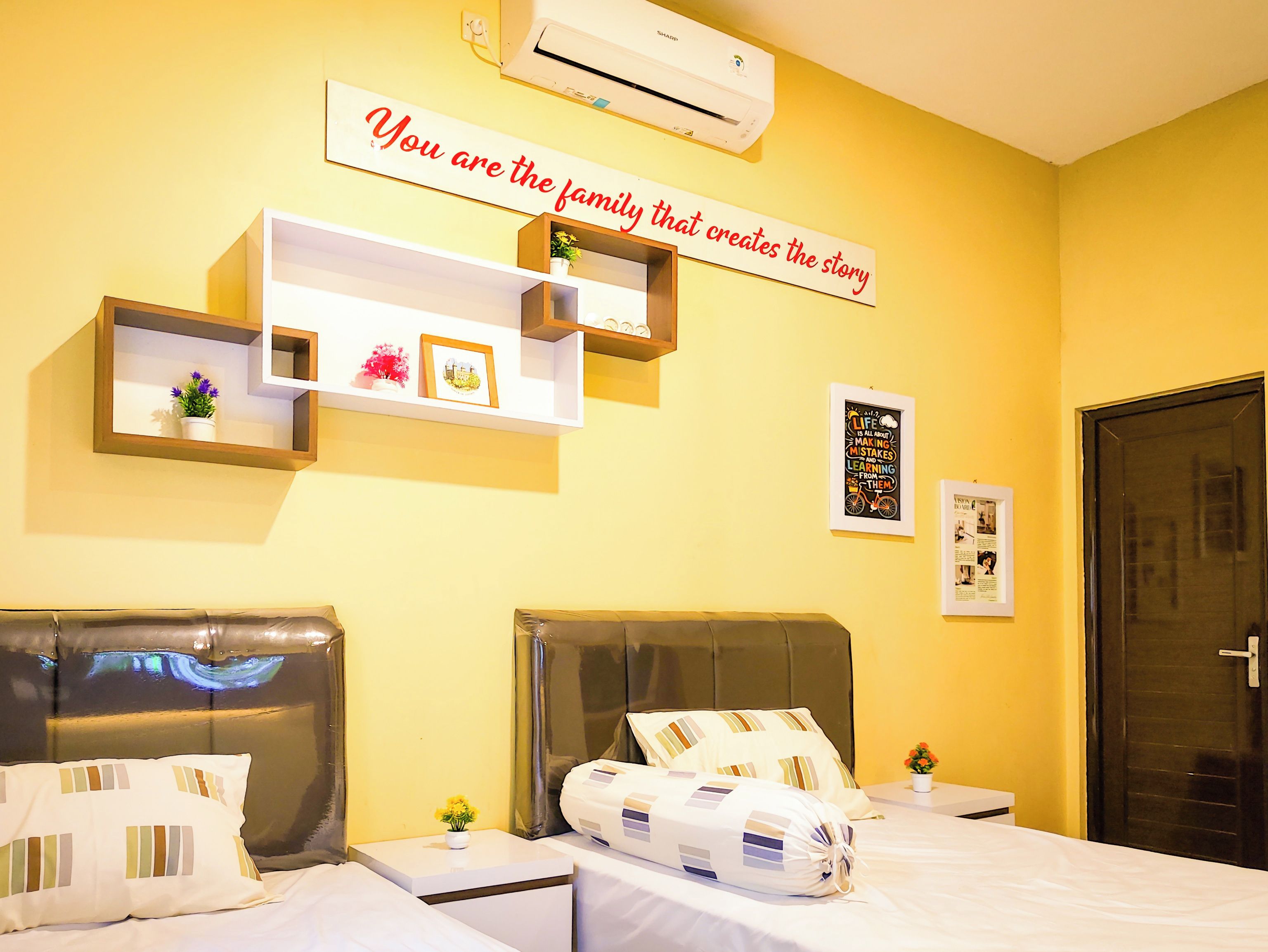 Quarto de hotel com cama, mesas de cabeceira, prateleira na parede com objetos decorativos, ar condicionado e uma porta.