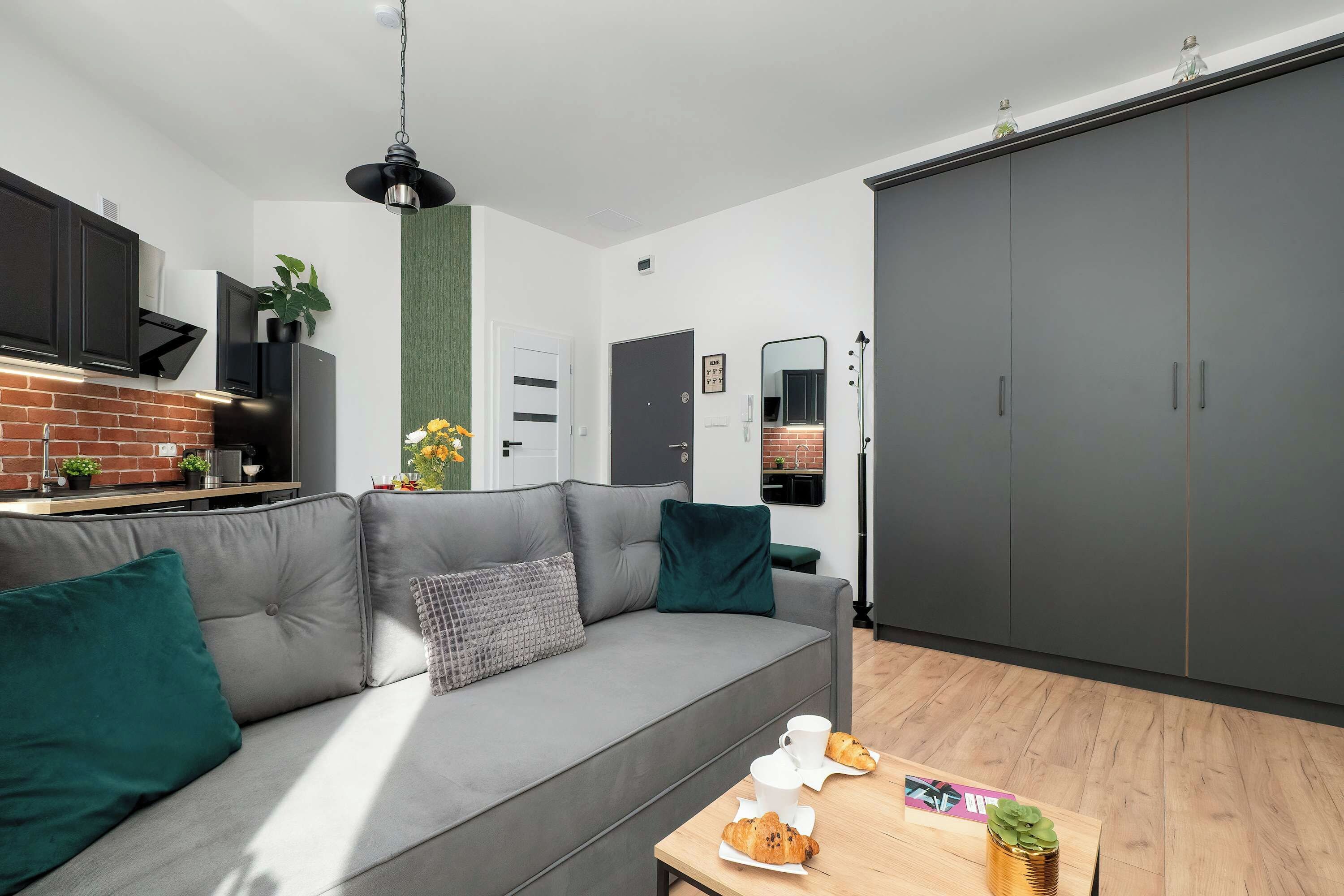 Et moderne køkken med et sort emfang, en grå sofa med grønne puder og et træspisebord med en croissant og en lille plante.