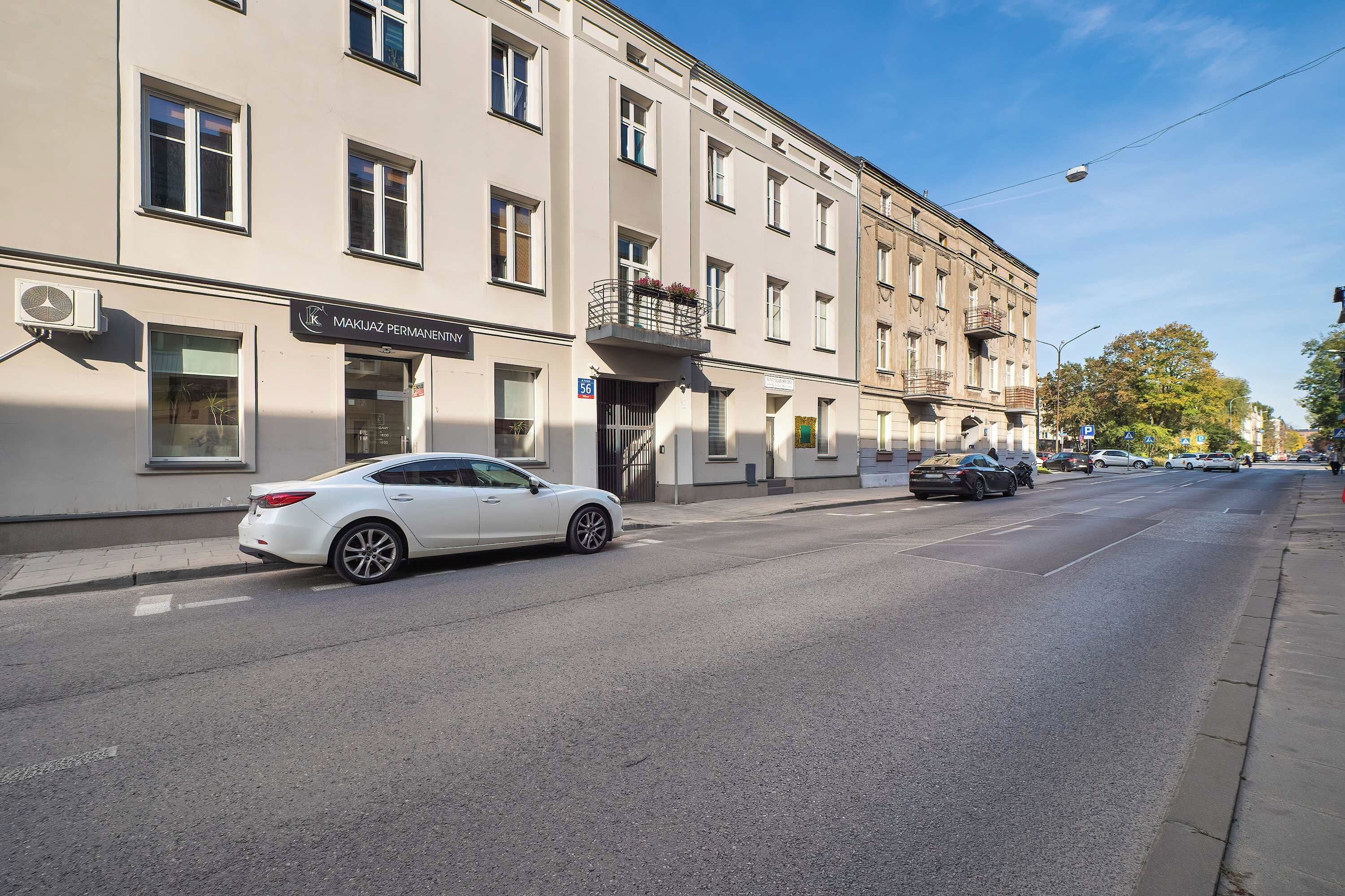 Straßenansicht mit einem weißen Auto, das vor einem Gebäude mit der Aufschrift 'MAKIJAŽ PERMANENTNY' geparkt ist.