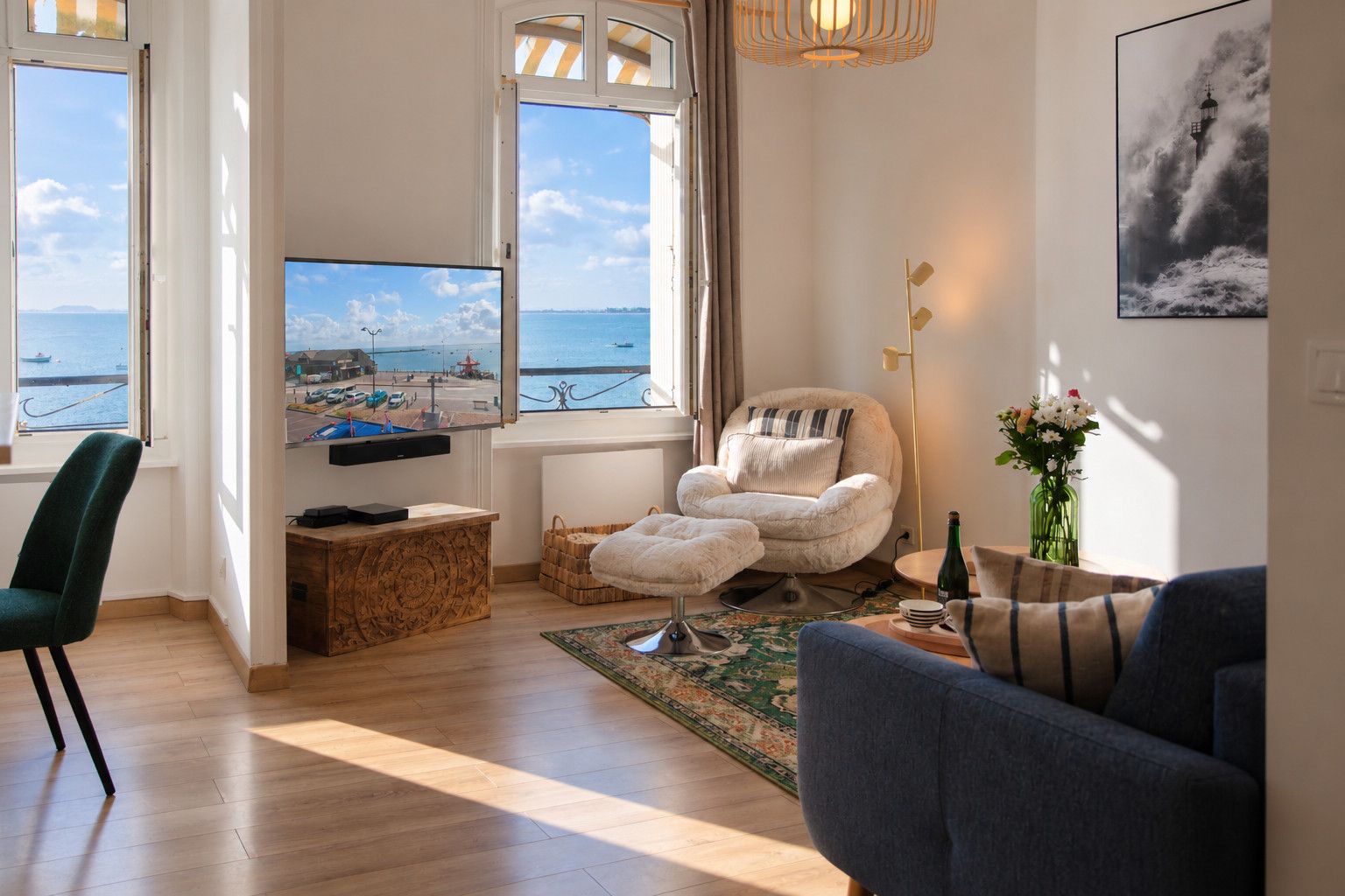 A living room with a television, a comfortable chair, a sofa, a wooden cabinet, and a view of the sea through large windows.