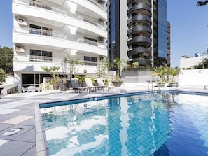 A swimming pool with lounge chairs, surrounded by a building with multiple balconies and a clear sky. - B Flat V (Porto Alegre)