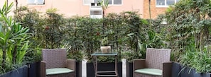 An outdoor seating area with wicker furniture and a glass table, surrounded by dense foliage and a building in the background. - B Flat V (Porto Alegre)