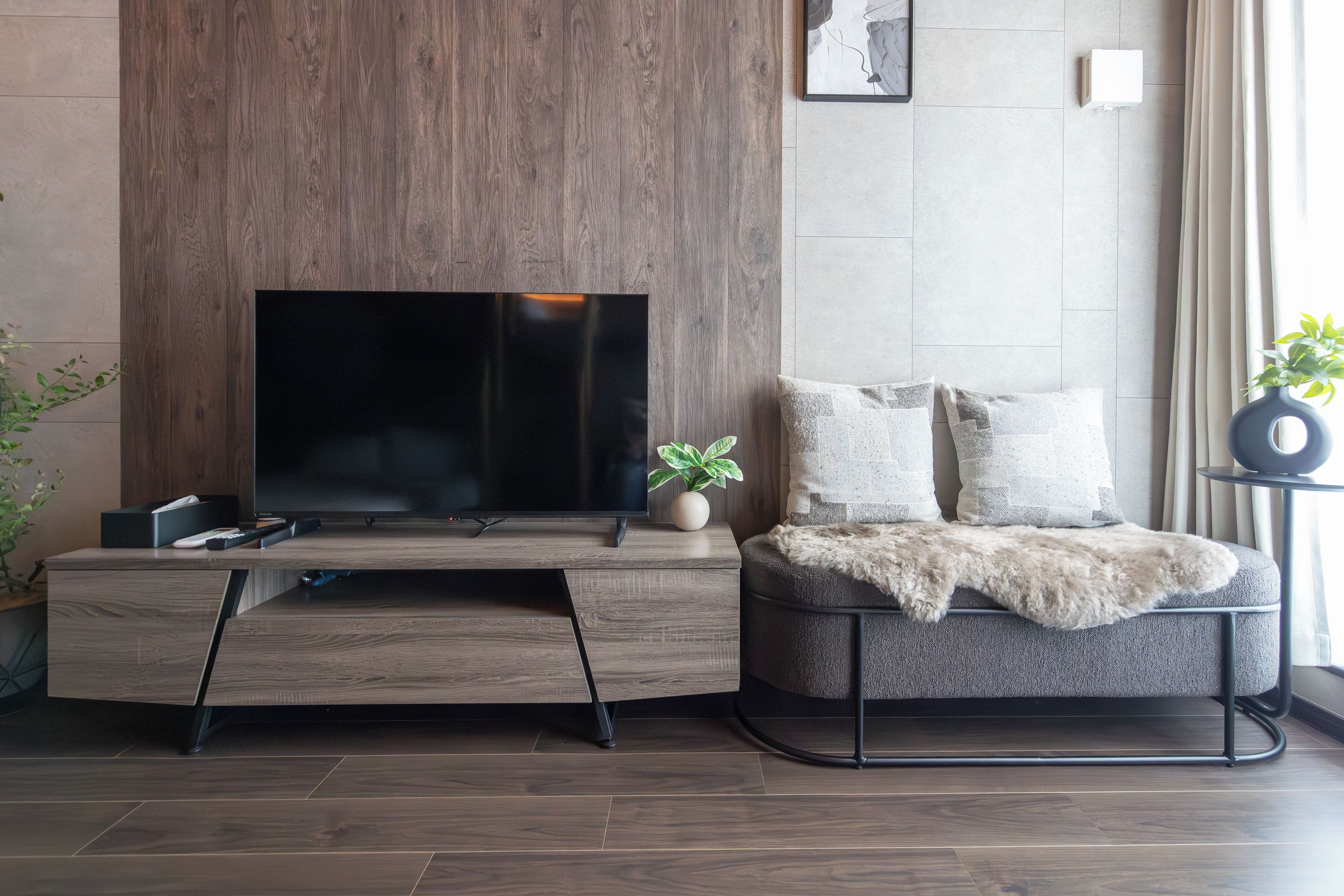 A modern living room with a flat-screen TV on a wooden stand, a grey sofa with a fur throw, and a small potted plant.