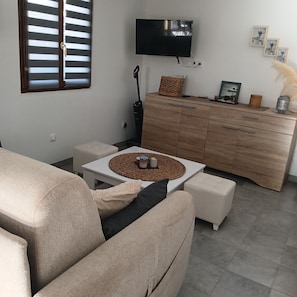 A living room with a sofa, a coffee table, a TV mounted on the wall, a wooden cabinet, and a window with blinds. - La Fontaine du Marais Vacation Rental, located near St. Valery-sur-Somme, with free parking (Pendé)