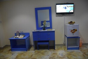 Una habitación con un escritorio azul, un espejo, un televisor y un mini refrigerador con fruta.