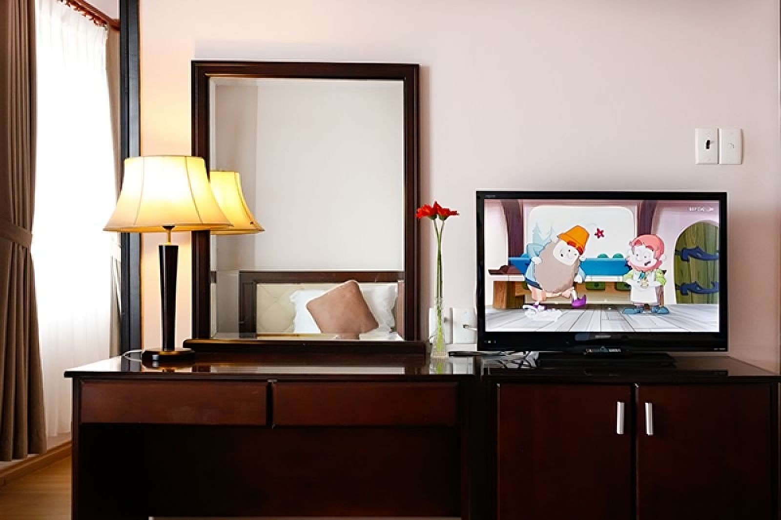 A hotel room with a television, a lamp, a mirror, and a vase with flowers.