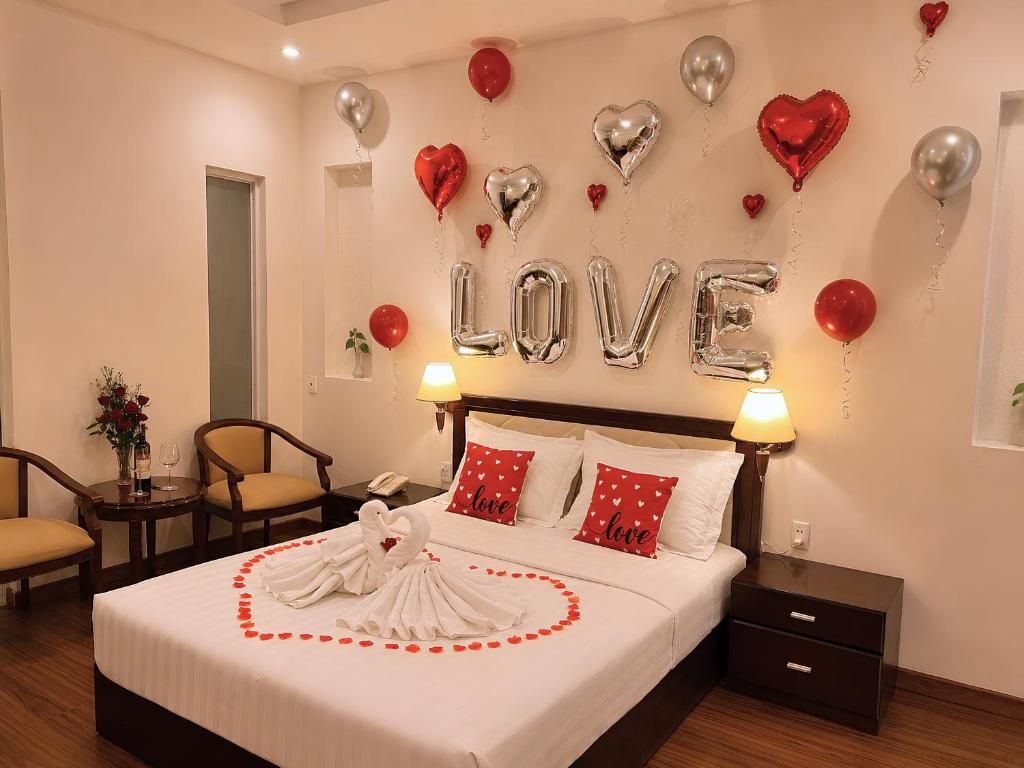 A hotel room with a bed, two bedside tables, a nightstand, a chair, a small table with a vase of flowers, and a wall with heart-shaped balloons and the word 'LOVE' spelled out in balloons.