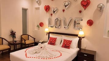 A hotel room with a bed, two bedside tables, a nightstand, a chair, a small table with a vase of flowers, and a wall with heart-shaped balloons and the word 'LOVE' spelled out in balloons.