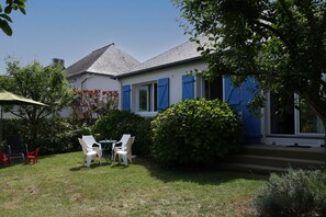 Uma casa com fachada branca e janelas azuis, um pequeno jardim com mesa e cadeiras, e uma árvore que oferece sombra.
