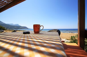 Ένα τραπέζι με ένα καρό τραπεζομάντιλο, ένα κόκκινο κούπα και ένα μαύρο αντικείμενο, με θέα μια παραλία και βουνά στο βάθος.