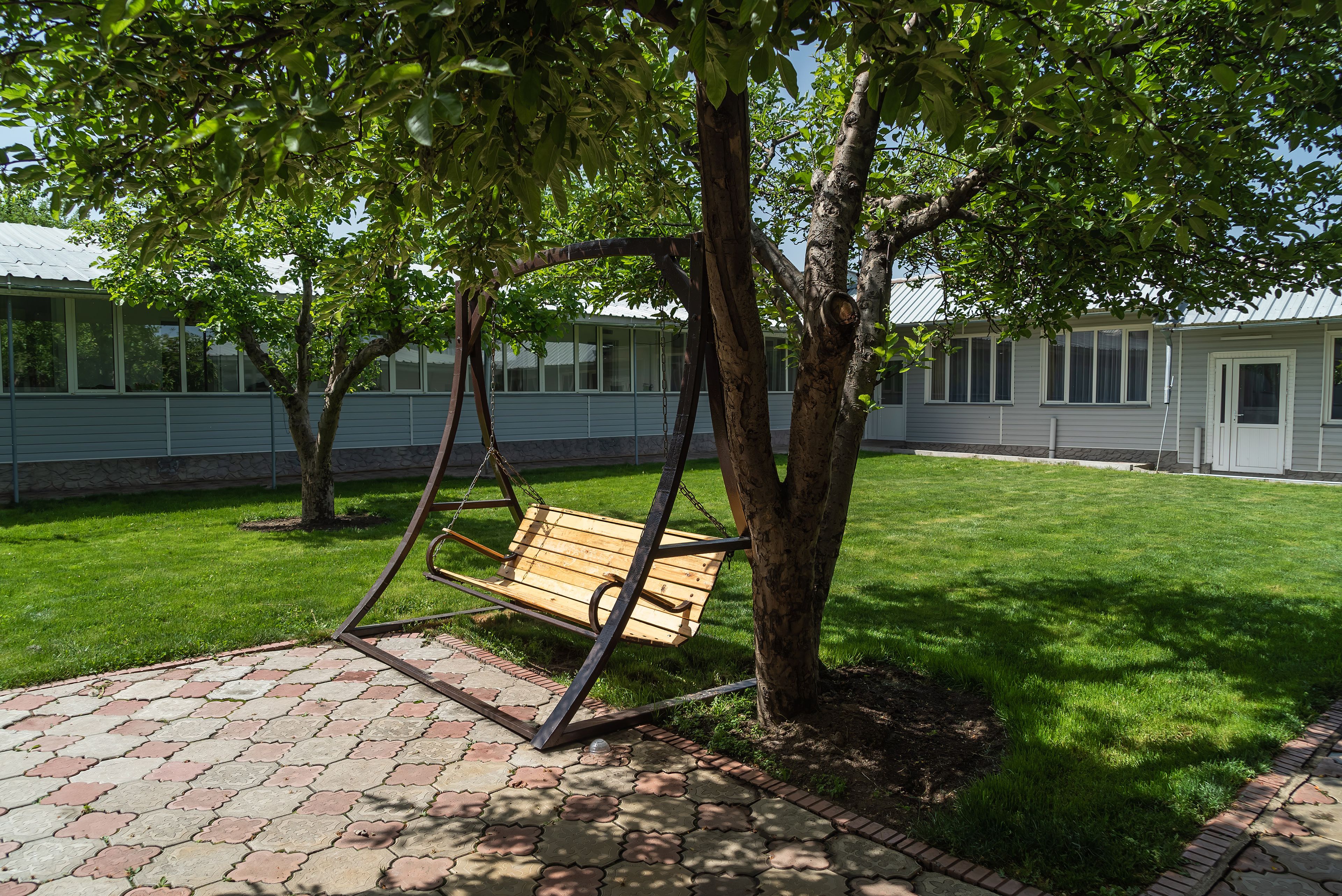 A swing set under a tree in a courtyard with a brick pathway and a building in the background.