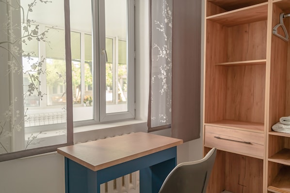 A room with a wooden desk, a chair, a window with a view of greenery, and a wooden wardrobe.