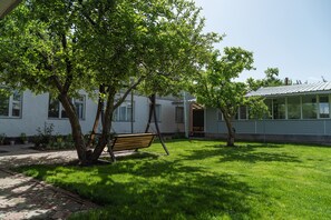 Une cour verdoyante avec un banc en bois, un arbre et un bâtiment doté de fenêtres et d’une porte.