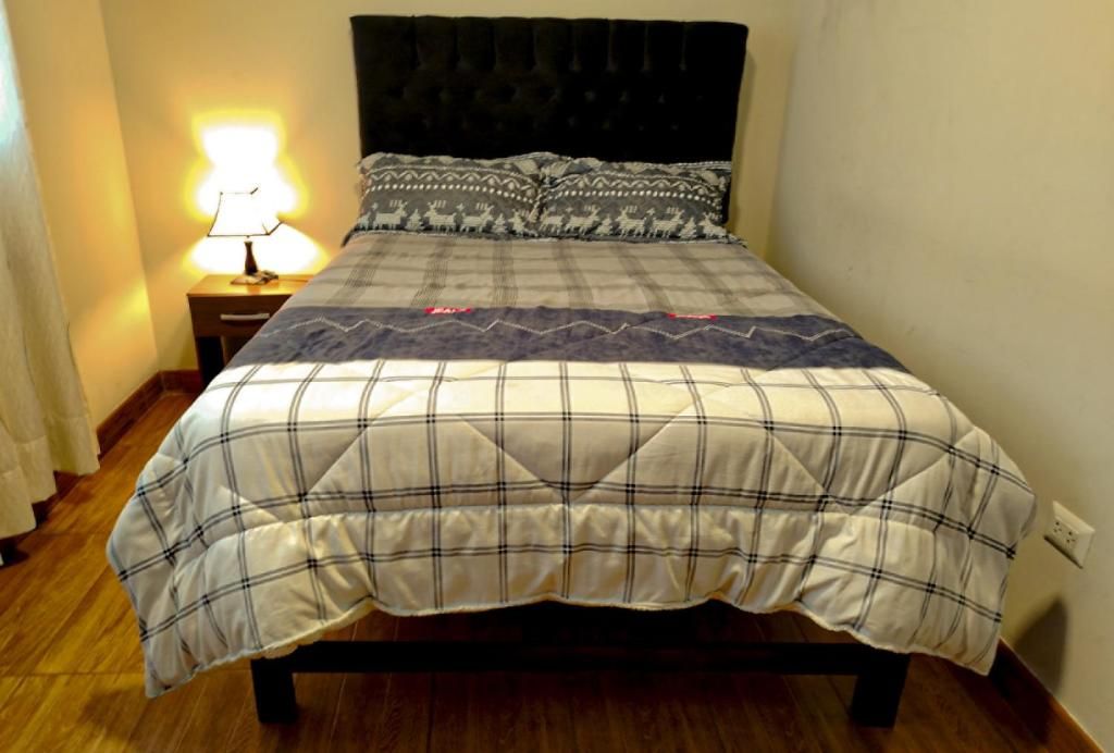 A bed with a plaid bedspread, a headboard, a bedside table with a lamp, and a wall socket.