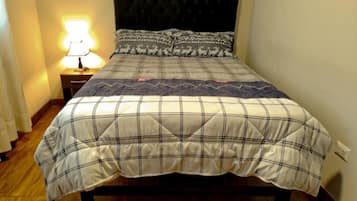 Una cama con colcha escocesa, cabecera, mesita de noche con lámpara y un enchufe en la pared.